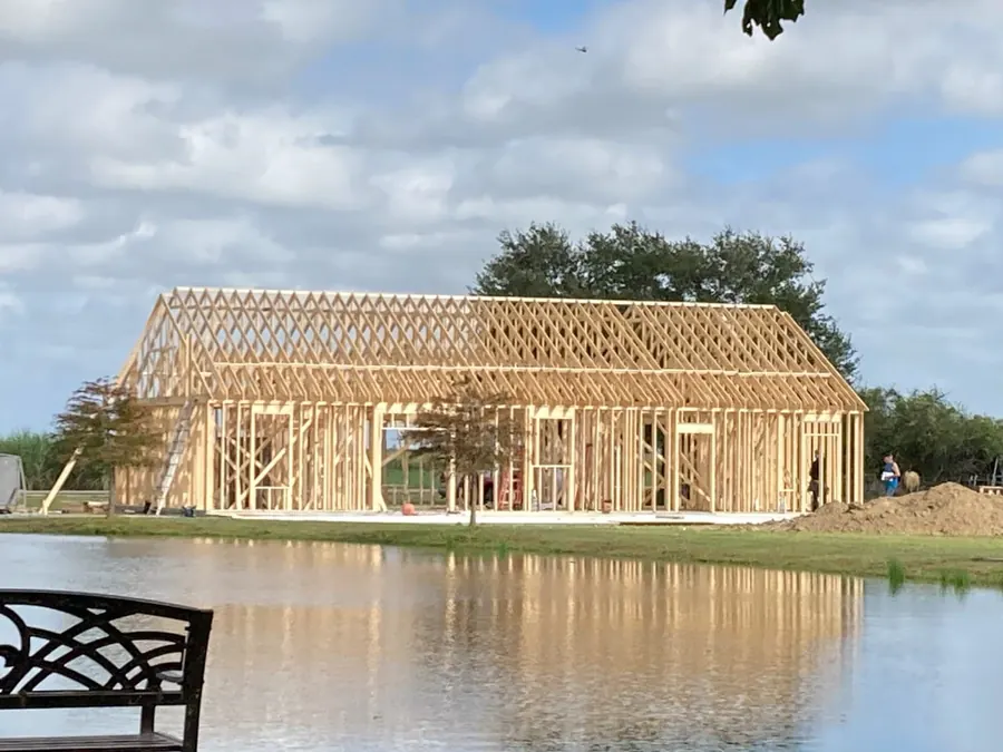 A home being framed beside a pond, with roof trusses set and blue sky above