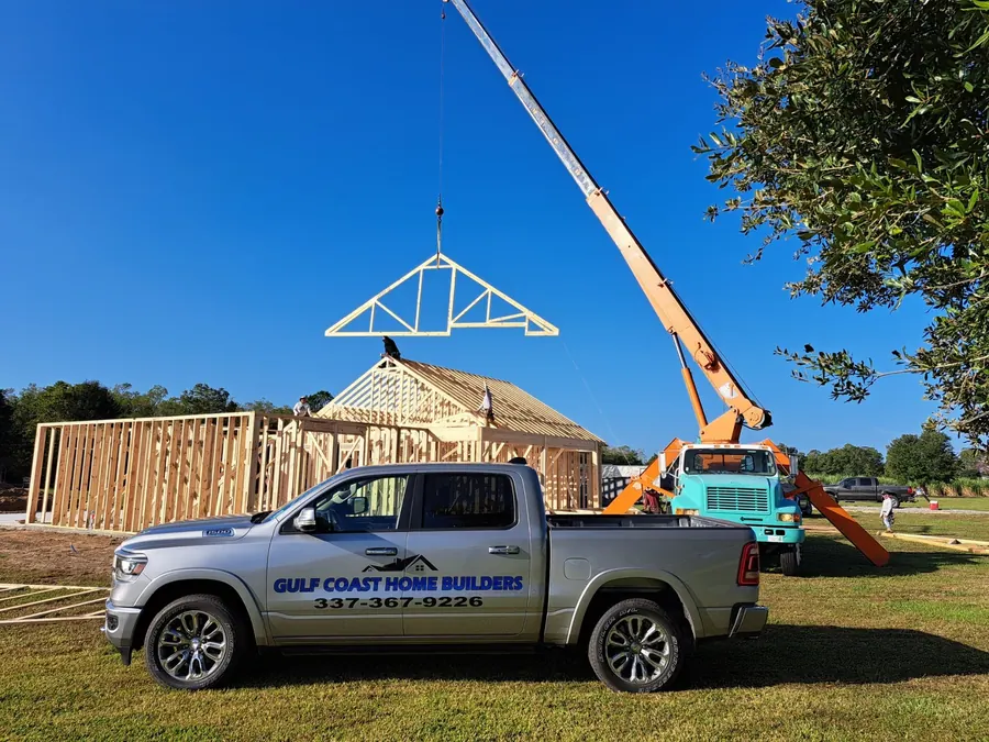 Crane setting a roof truss on a home under construction, with a Gulf Coast Home Builders truck in front