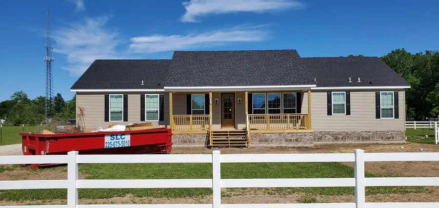 Tan home with black shutters, a covered front porch with wood rails and a white fence